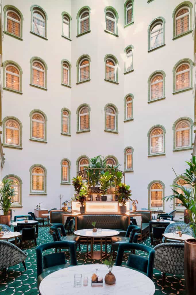 Courtyard lounge of the Párisi Udvar Hotel under a glass roof, white walls with rows of arched windows rising above green velvet armchairs, round tables, plants and a bar