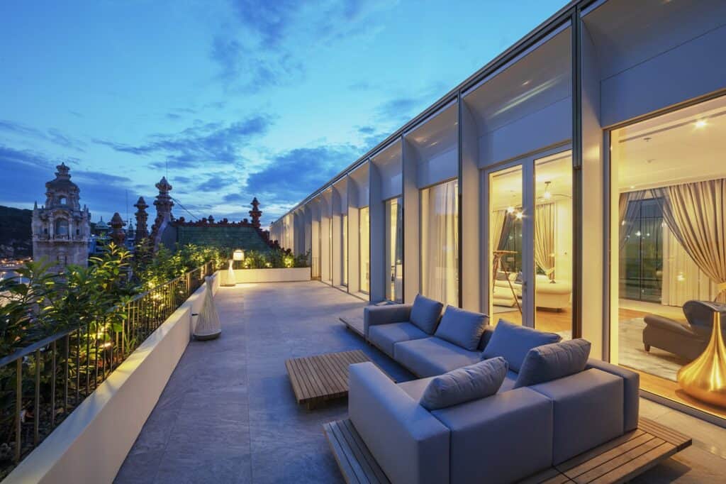 Rooftop terrace on the new top floor of the Párisi Udvar at dusk, grey sofas and a timber deck along a glazed wall, looking out to the turrets and spires of Budapest under a cloudy blue sky