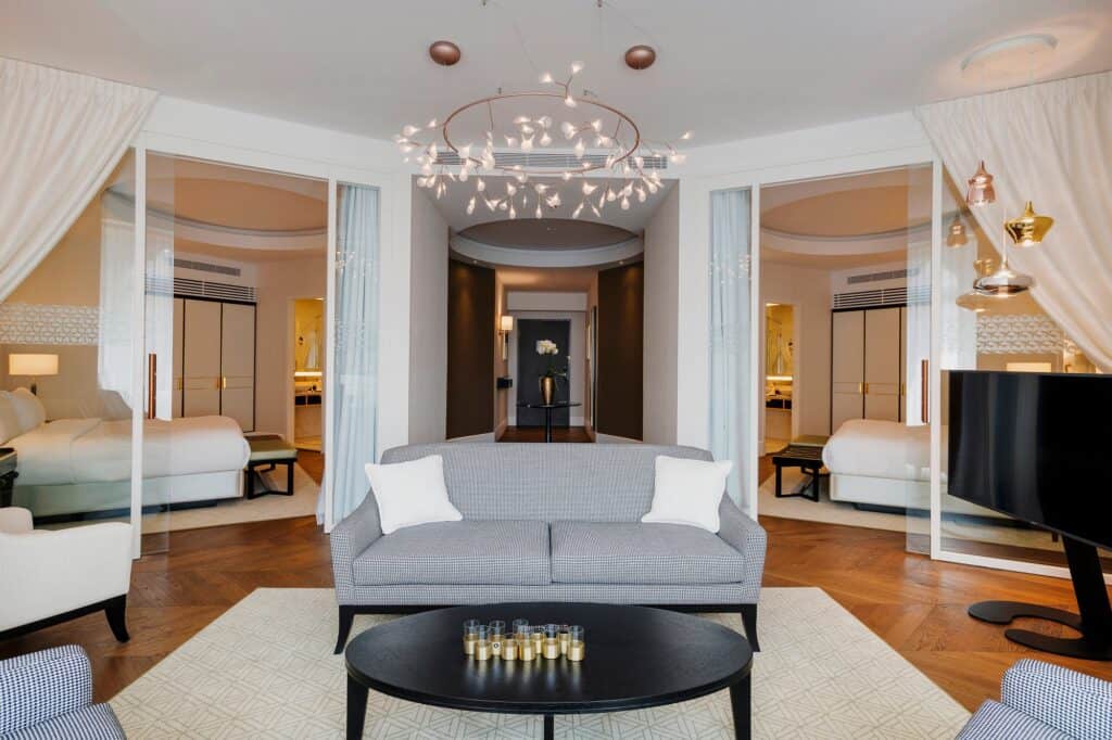 Suite at the Párisi Udvar Hotel with a pale grey sofa and dark oval coffee table on a cream rug, a chandelier of glass globes and glazed partitions opening to a bedroom on each side