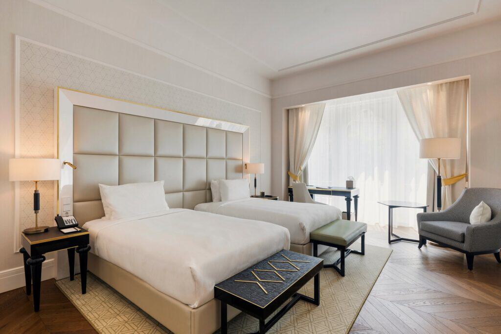 Guest room with two white beds against a tall quilted grey headboard, dark benches at their feet, a grey armchair by curtained windows and a herringbone timber floor