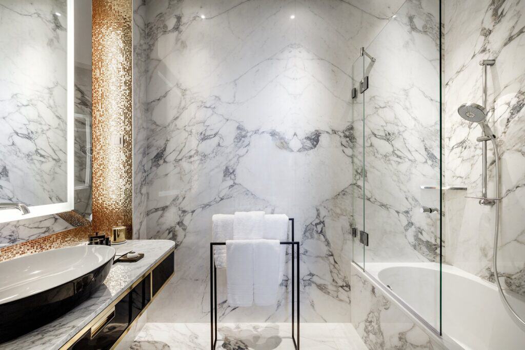 Marble bathroom with a dark vanity and stone basin, a walk-in glass shower and a bathtub beyond a glass screen, with a gold mosaic panel beside the mirror