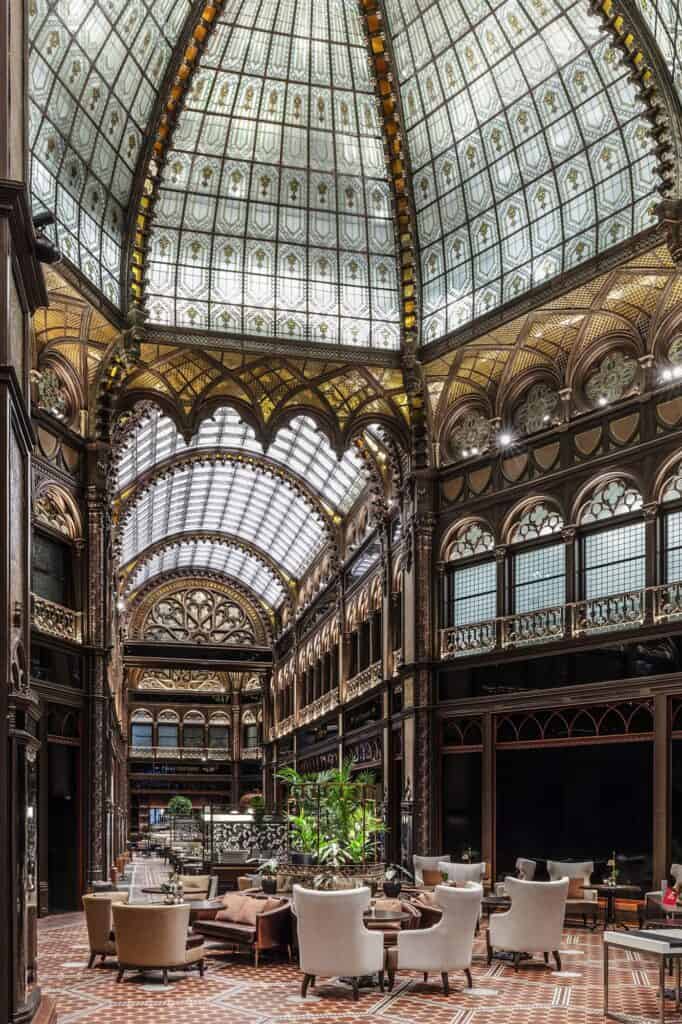 Ground level of the Paris Court arcade set with white armchairs and low tables beneath the glass dome, carved dark timber galleries and stained glass windows on three sides