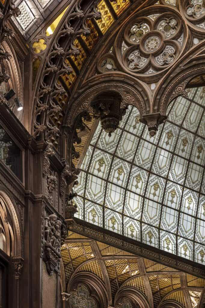Close view of carved Neo-Gothic tracery and timber columns in the Párisi Udvar, framing a panel of patterned leaded glass and a glimpse of the vaulted roof