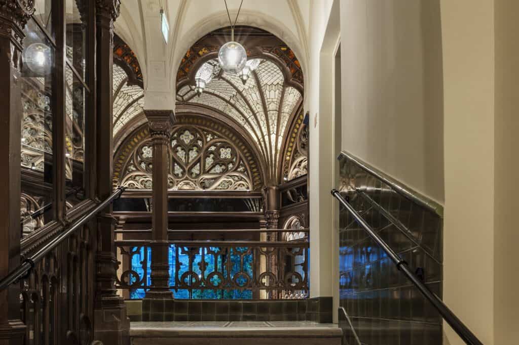 View from a stairwell with a black glass balustrade into the Paris Court arcade, a carved stained glass arch, a hanging lantern and a blue-lit gallery panel