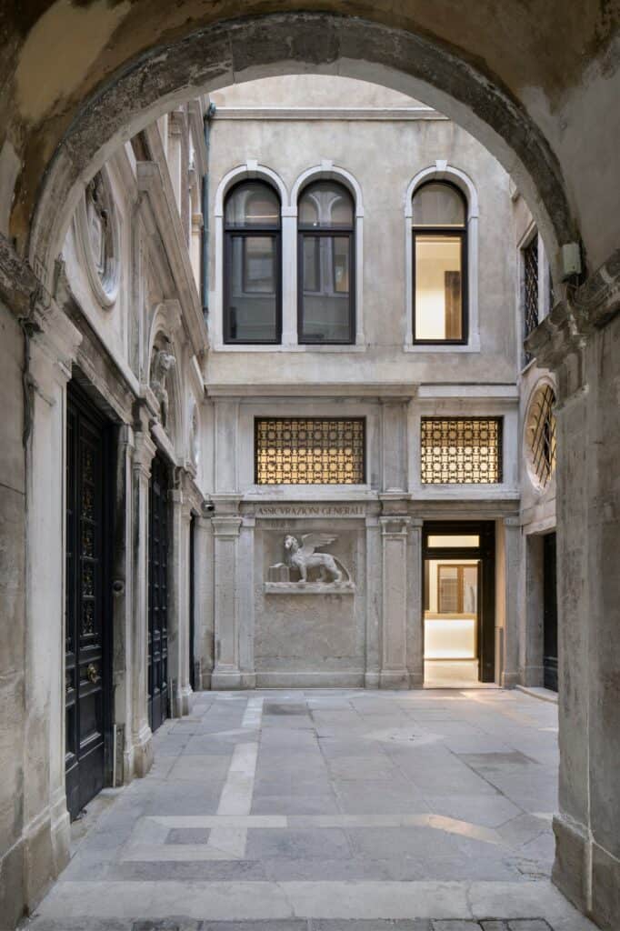 View through a stone archway into a courtyard of the Procuratie Vecchie, a winged lion of St Mark relief set below ornate window grilles, with arched windows above and tall dark doors along the stone paving