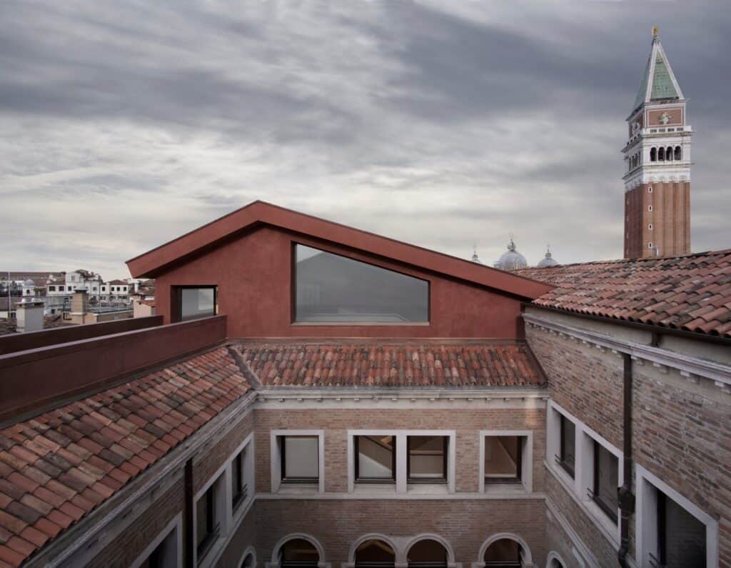 New red-plastered roof pavilion with a wide window rising above the terracotta roofs and brick inner courtyard of the Procuratie Vecchie, the St Mark's campanile and basilica domes beyond under a cloudy sky