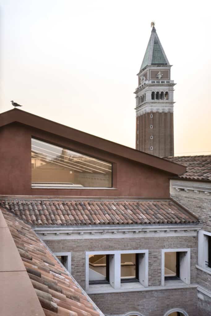 Gabled red roof pavilion of the Procuratie Vecchie with a long window above terracotta tiles and brick courtyard walls, the St Mark's campanile glowing behind at dusk and a gull perched on the roof