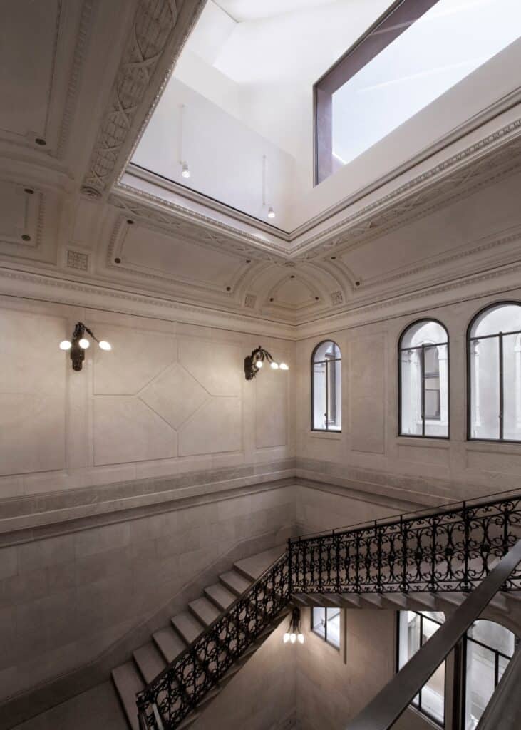 Historic staircase in the Procuratie Vecchie with an ornate wrought-iron balustrade, stucco ceiling mouldings and brass wall lamps beneath a new skylight and arched windows