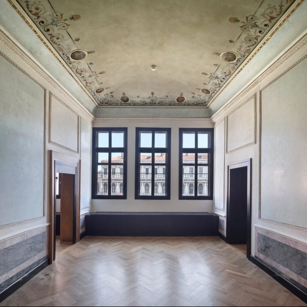 Restored room in the Procuratie Vecchie with a painted ceiling of scrolling garlands and medallions, pale panelled walls, a herringbone parquet floor and three dark-framed windows facing an arcaded facade
