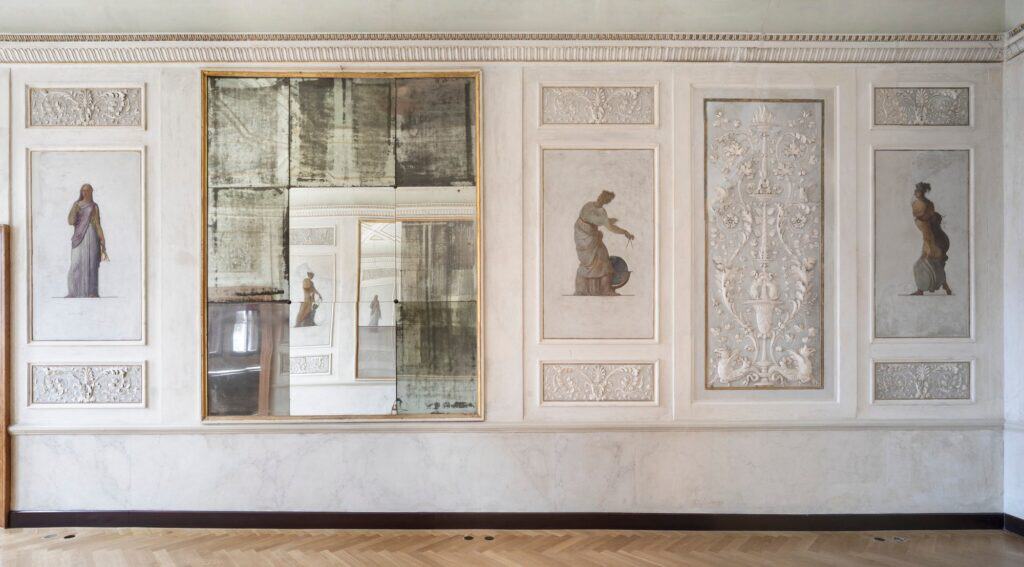 Restored wall in the Procuratie Vecchie lined with grisaille panels of classical female figures and carved ornament either side of an antique foxed mirror, above a herringbone parquet floor