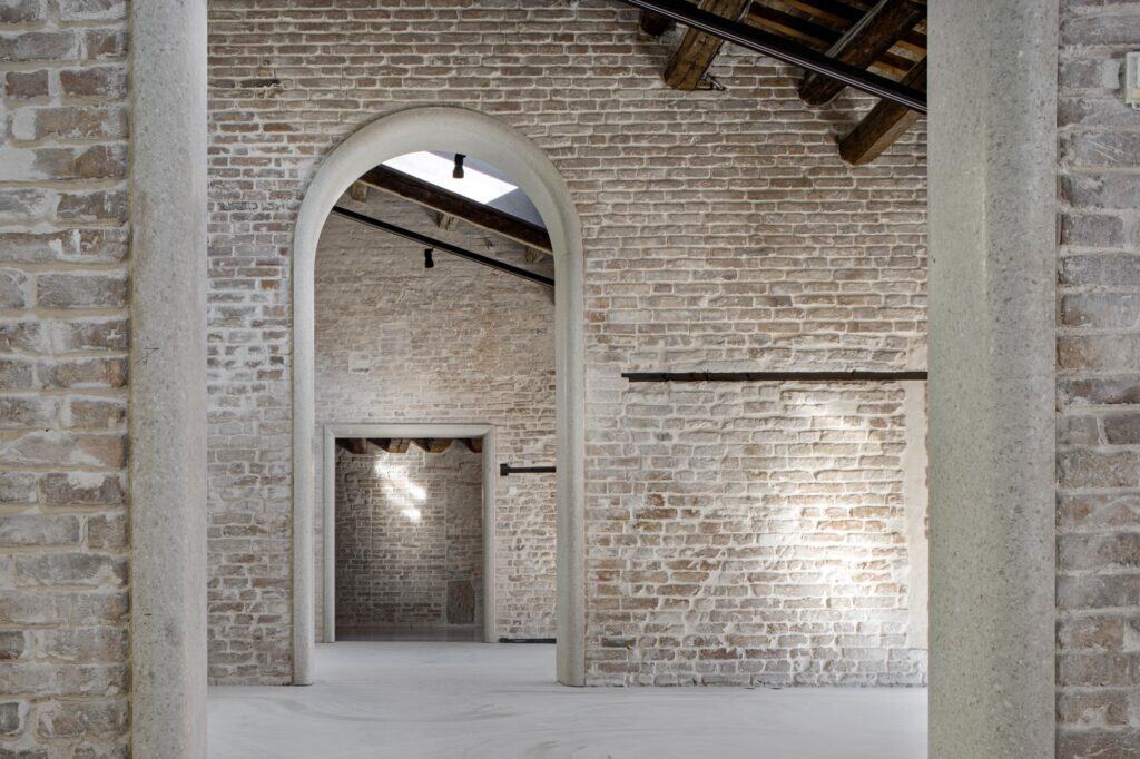 Room in the Procuratie Vecchie with uncovered brick walls and dark timber roof beams, a pale stone arch framing a doorway into a further brick room lit from above