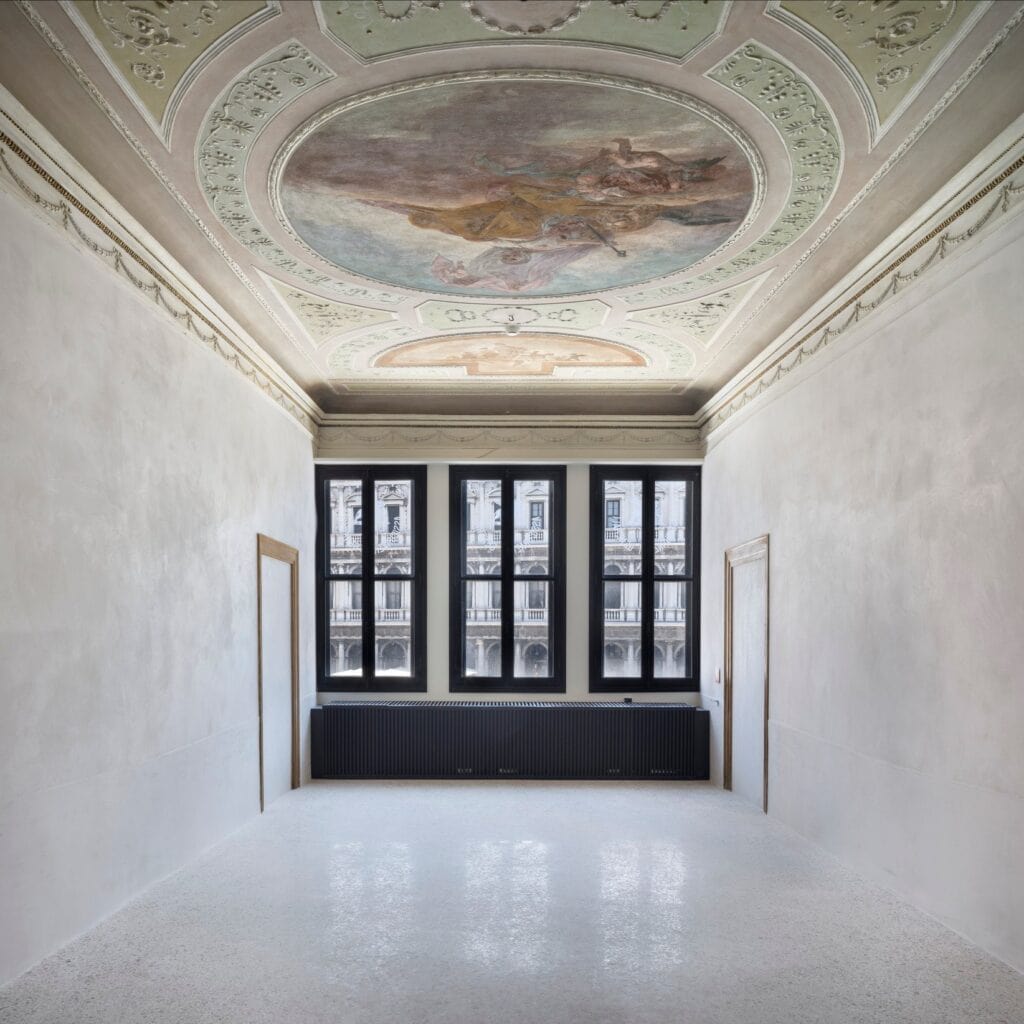 Restored room in the Procuratie Vecchie beneath a frescoed ceiling with an oval painting of figures in the sky framed by pale green stucco, white walls, a terrazzo floor and three windows facing an arcaded facade