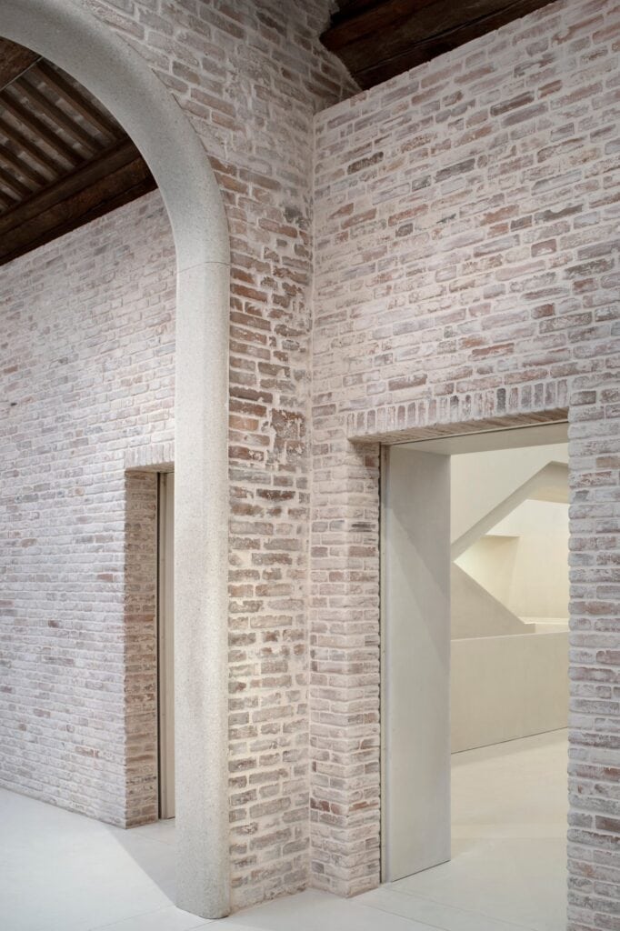 Tall pale stone arch set into uncovered brick walls of the Procuratie Vecchie beneath dark timber beams, opening into a white space with a glimpse of the new staircase