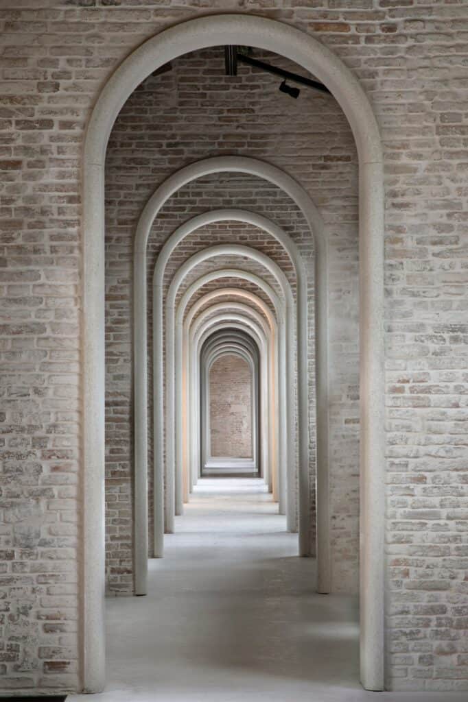 Enfilade of slender stone arches receding through the brick walls of the third floor of the Procuratie Vecchie, a long perspective down a pale floor to a brick end wall