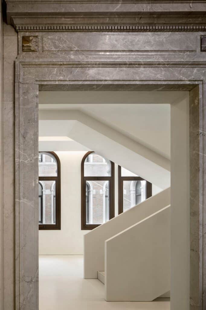 Grey marble door frame in the Procuratie Vecchie opening onto a bright white stair hall, the angular flights of the new staircase rising past tall arched windows