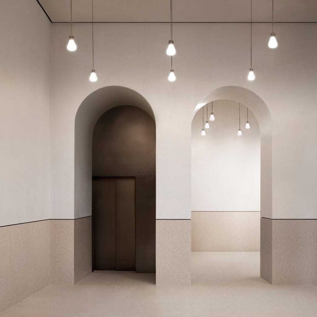 Two arched openings in a pale plastered entrance hall of the Procuratie Vecchie, one framing a bronze-toned lift door, with clusters of bare-bulb pendants above a speckled stone dado
