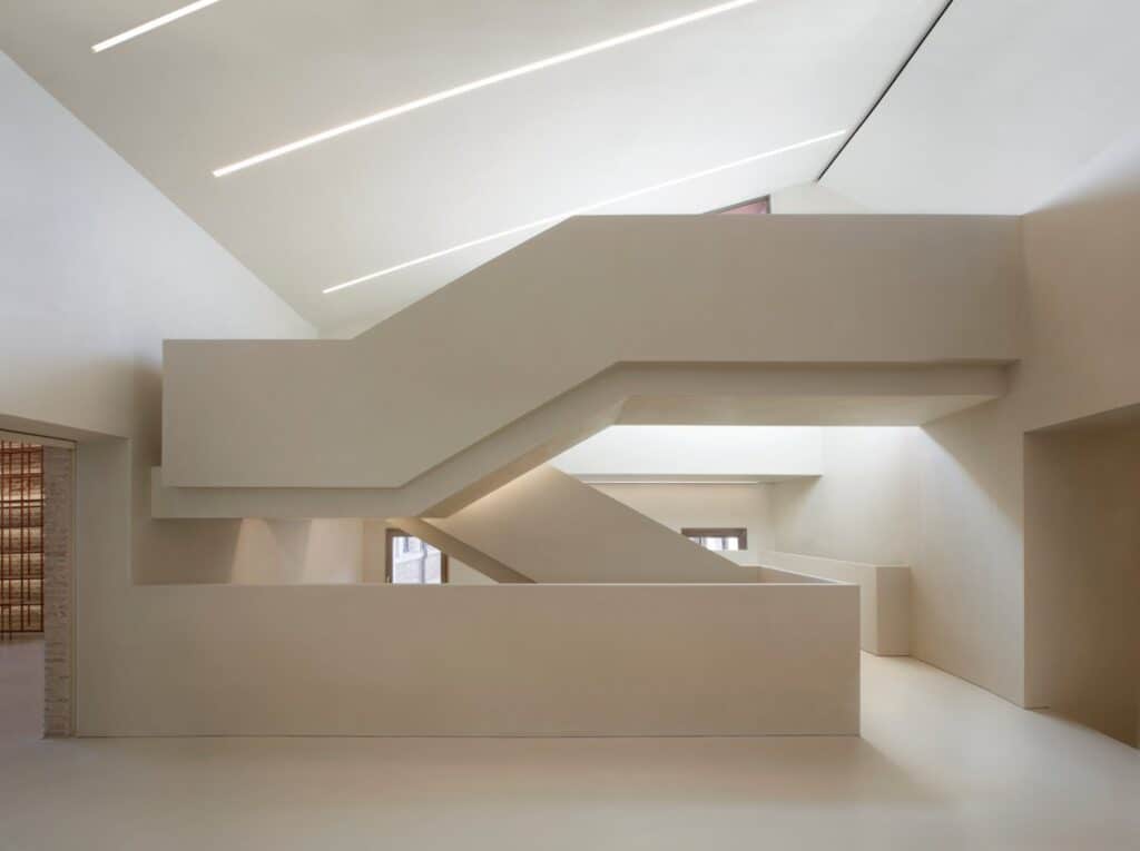 Angular white flights of the new staircase in the Procuratie Vecchie interlocking beneath a sloping ceiling lit by linear light strips