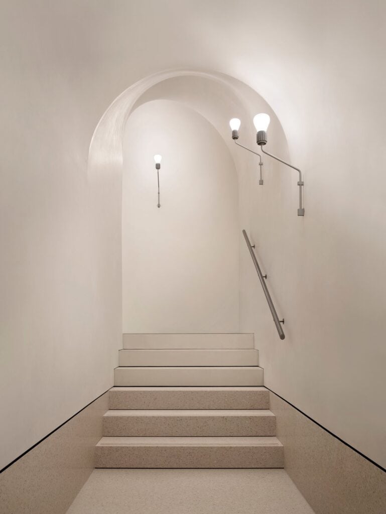 Speckled stone steps rising beneath a plastered arch in the Procuratie Vecchie, lit by slender wall lamps with a metal handrail fixed to the wall