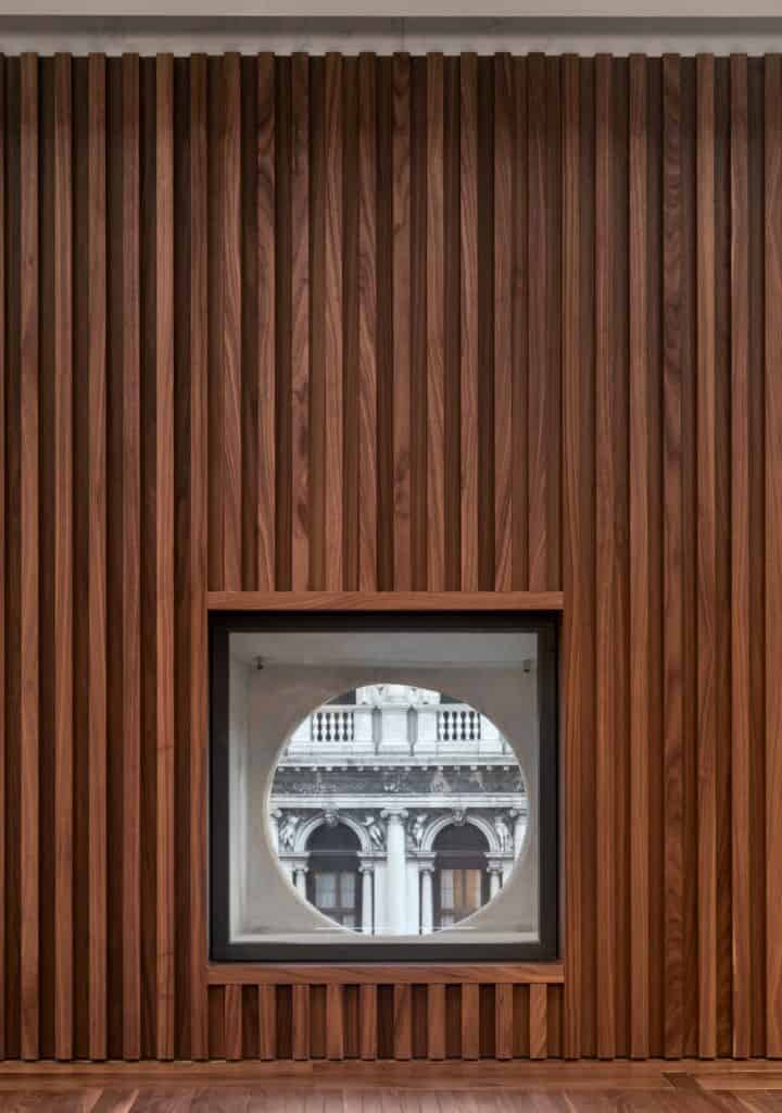 Walnut-toned vertical timber slats lining the auditorium of the Procuratie Vecchie around a deep window whose circular opening frames a glimpse of an ornate classical facade