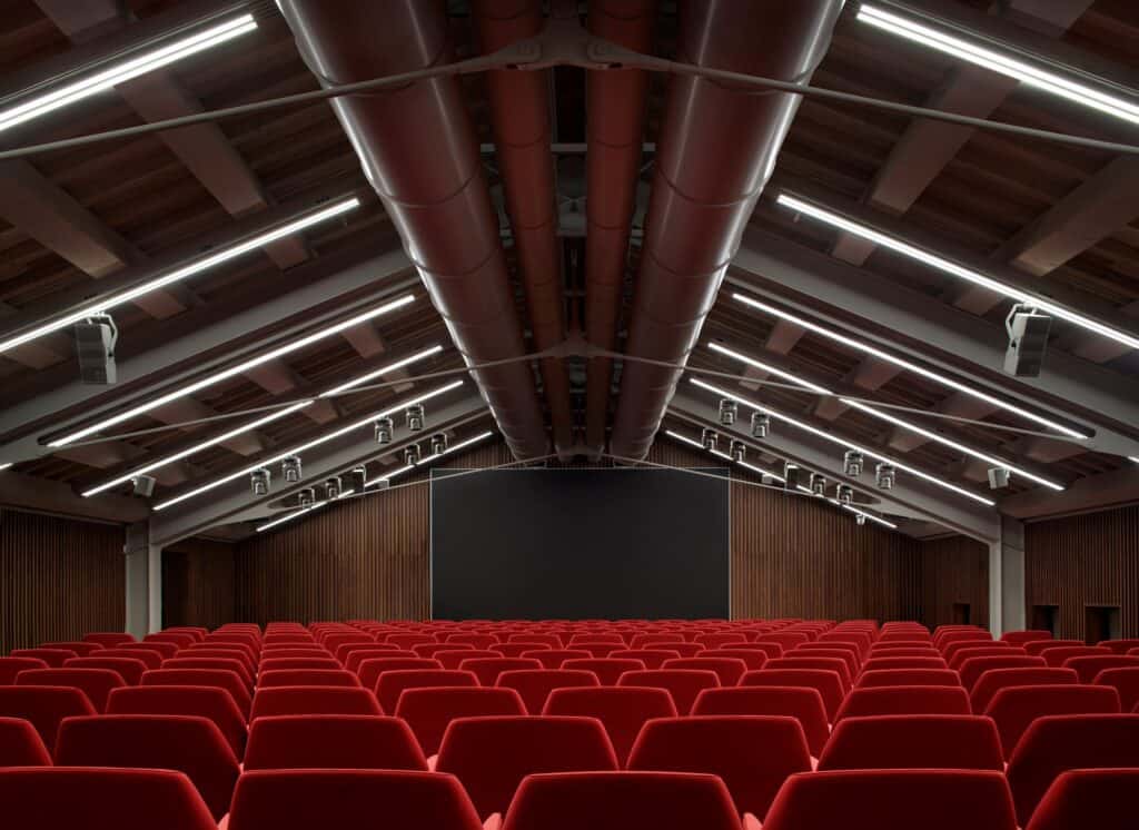 Rows of red seats in the auditorium of the Procuratie Vecchie beneath exposed timber roof trusses, with linear lights, red ducts and timber-panelled walls around a dark screen