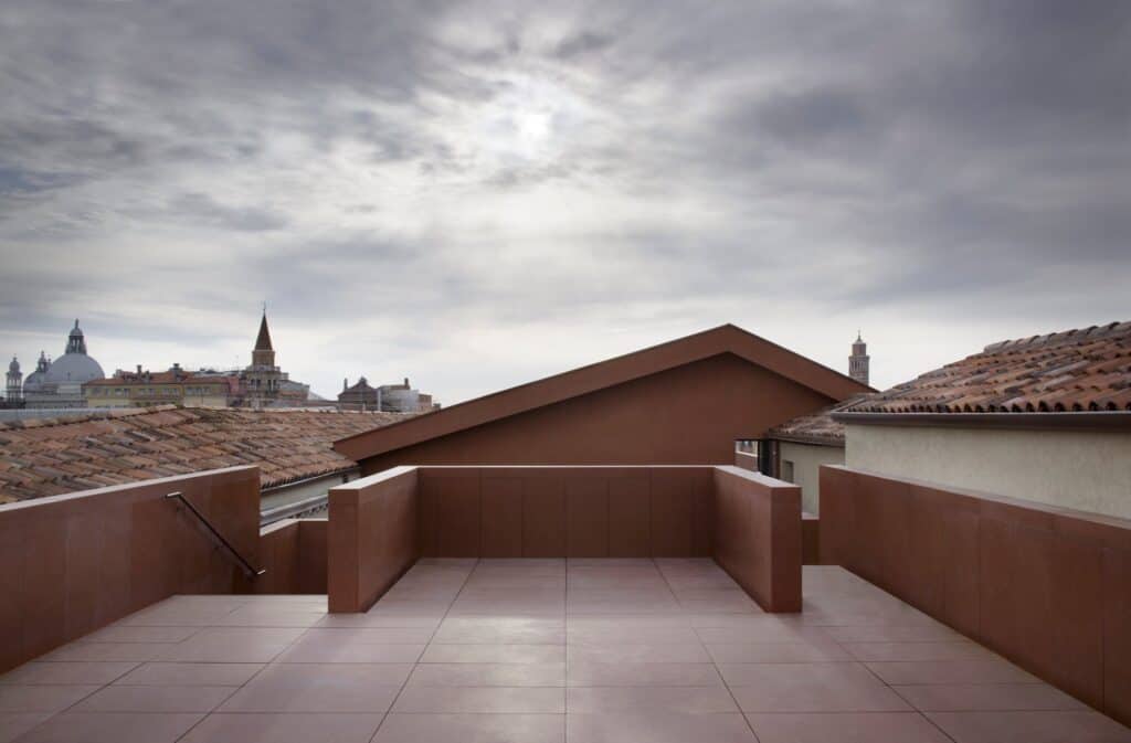 Roof terrace of the Procuratie Vecchie paved in terracotta-toned tiles with red parapets and the gabled pavilion, Venetian domes and bell towers on the skyline under a cloudy sky