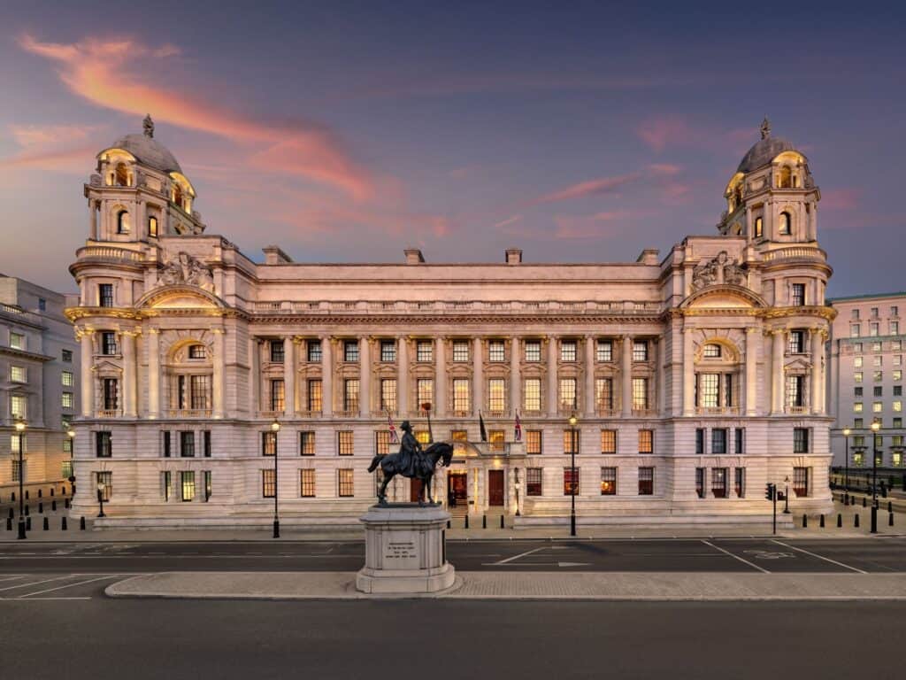 Whitehall façade of the Old War Office at dusk, now Raffles London at The OWO, its Edwardian Baroque stone front lit warm gold between twin domed corner towers, with an equestrian statue on a plinth in the foreground under a pink sky