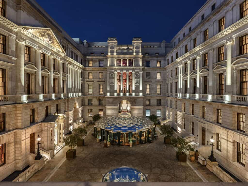 Central courtyard of The OWO at night, floodlit stone façades rising on three sides around a glass pavilion with a blue patterned canopy, potted olive trees and lanterns along the paving