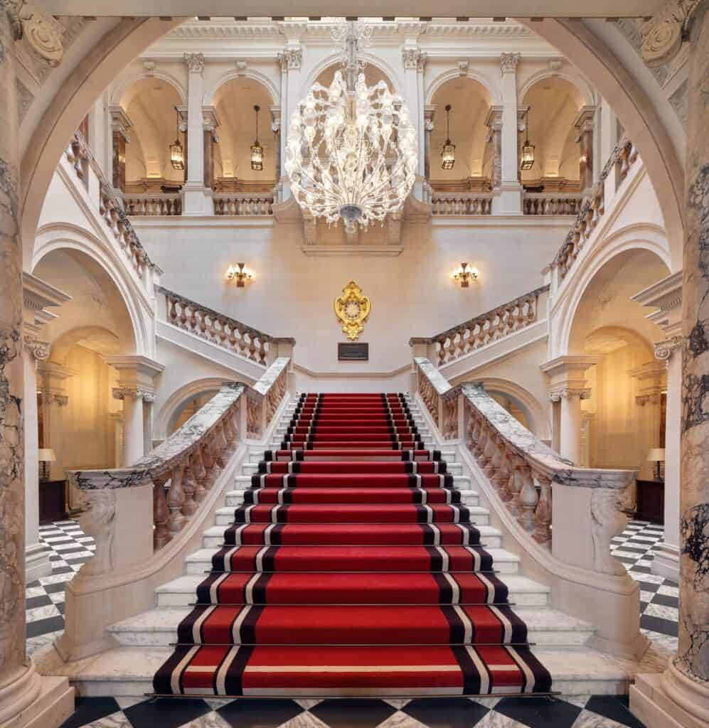 Grand marble staircase of Raffles London at The OWO, a red carpet with black borders rising to a landing where the flights split, alabaster balustrades and arched galleries around a tiered crystal chandelier