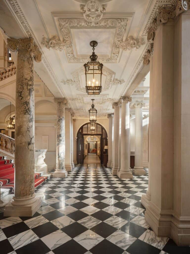 Colonnaded hall at Raffles London with a black and white chequerboard marble floor, fluted stone columns and an ornate plaster ceiling hung with brass lanterns leading to an arched doorway