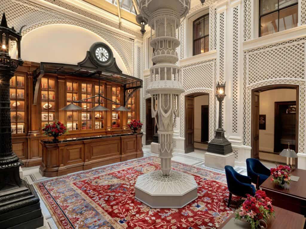 Concierge hall at Raffles London at The OWO, a carved white openwork column on an octagonal base standing on a red patterned rug, before a mahogany cabinet crowned by a clock and walls of white lattice panels