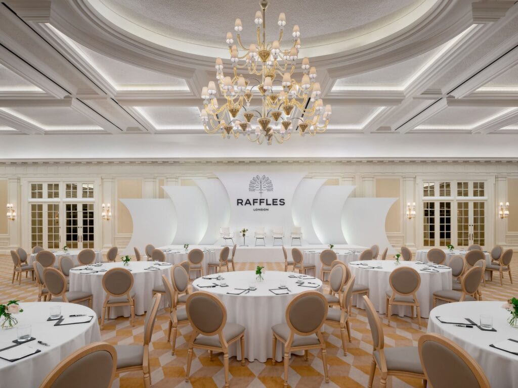 Whitehall Ballroom set for a banquet, round tables with white cloths and cream chairs under a coffered ceiling with a large gold and crystal chandelier, and a white stage with the Raffles crest