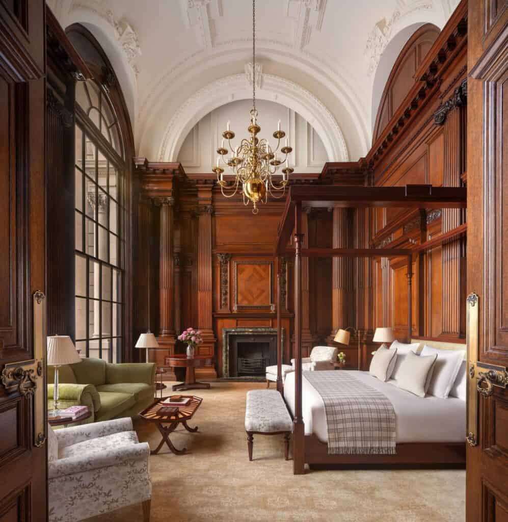 Panelled bedroom suite at Raffles London with a dark timber four-poster bed and plaid throw, a barrel-vaulted white ceiling with a brass chandelier, a marble fireplace and tall arched windows