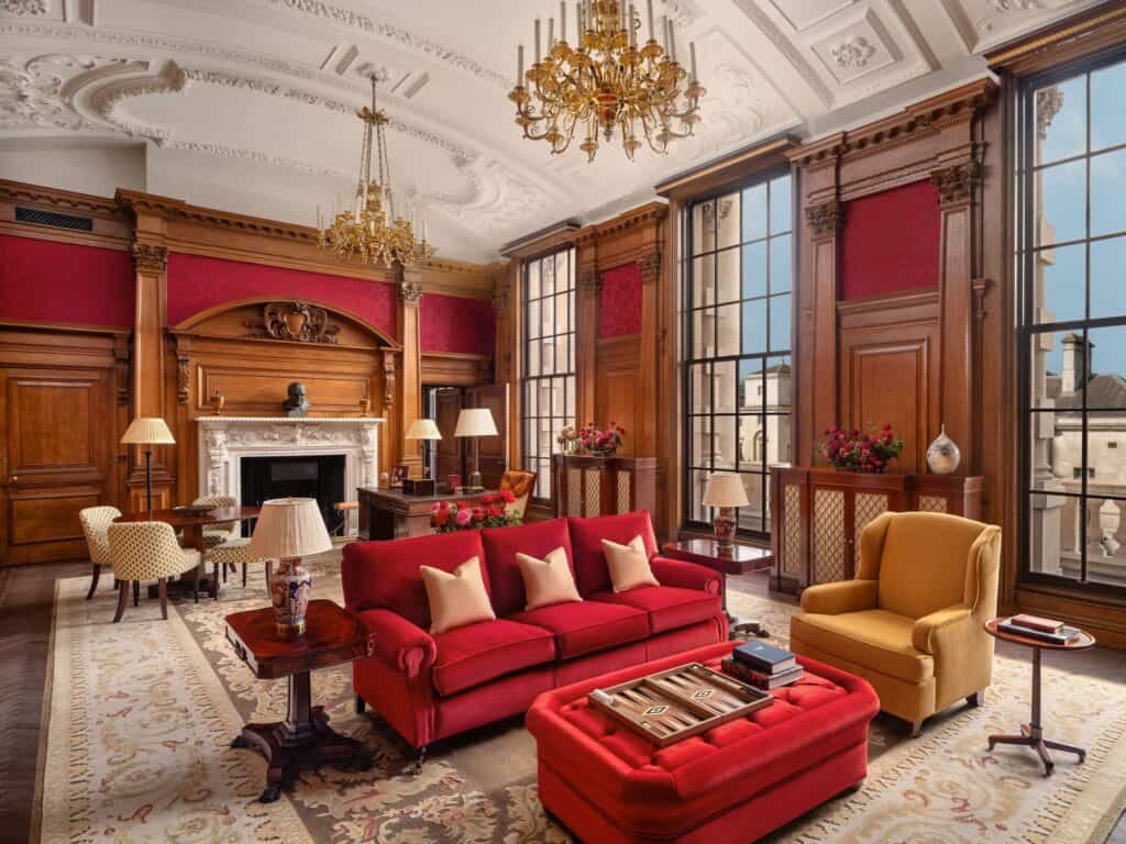 Haldane heritage suite at Raffles London at The OWO, red damask panels set into oak panelling, a red velvet sofa and ottoman with a backgammon board, a mustard armchair and tall windows
