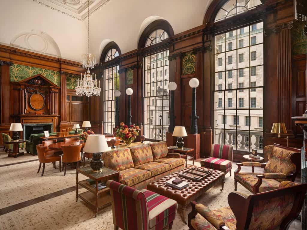 Churchill suite living room, the former Army Council Room, with dark oak panelling and a green damask frieze, tall arched windows, crystal chandeliers, a patterned sofa and tufted leather ottoman before a carved fireplace