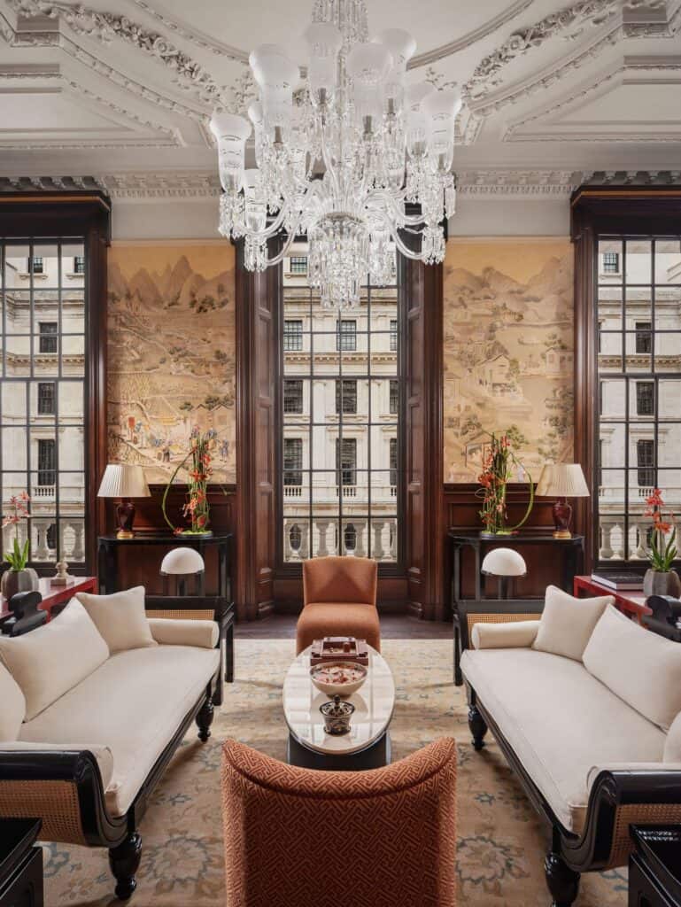 Raffles Suite sitting room with a pair of white daybeds facing across a round table, a crystal chandelier under an ornate cornice and hand-painted landscape panels between tall sash windows