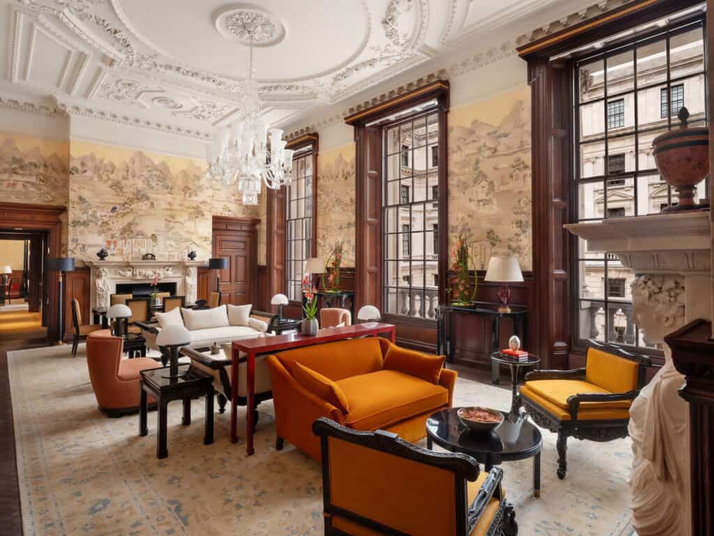 Raffles Suite drawing room with scenic painted wall panels, orange velvet armchairs and black lacquered tables, an ornamental plaster ceiling with a chandelier and a carved marble fireplace