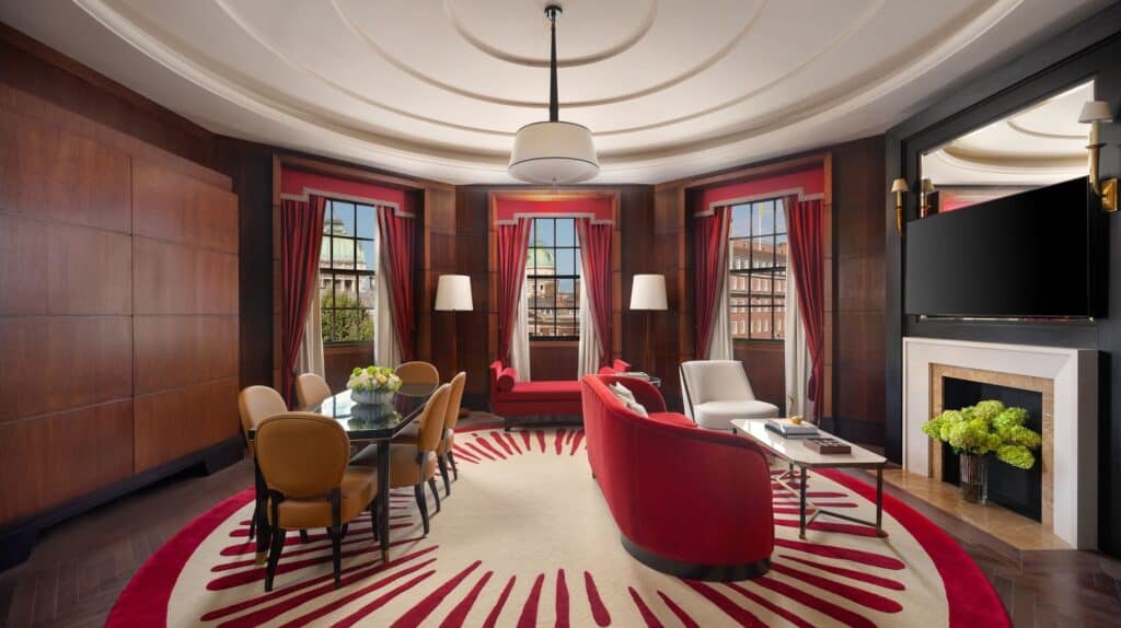 Art Deco Atkins corner suite with a curved timber-panelled wall, a round red and cream sunburst rug, a red curved sofa and a dining table before three red-curtained windows