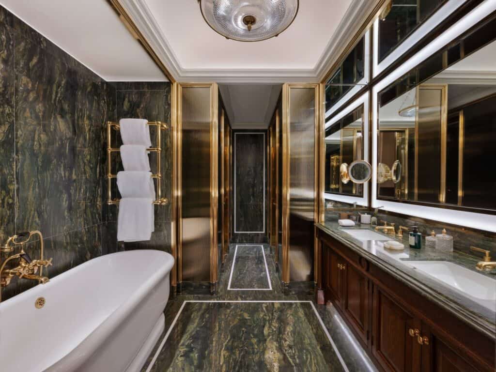 Marble bathroom at Raffles London with a freestanding white bathtub, dark green veined marble walls and floor, brass taps and a towel rail, and a long vanity with illuminated mirrors