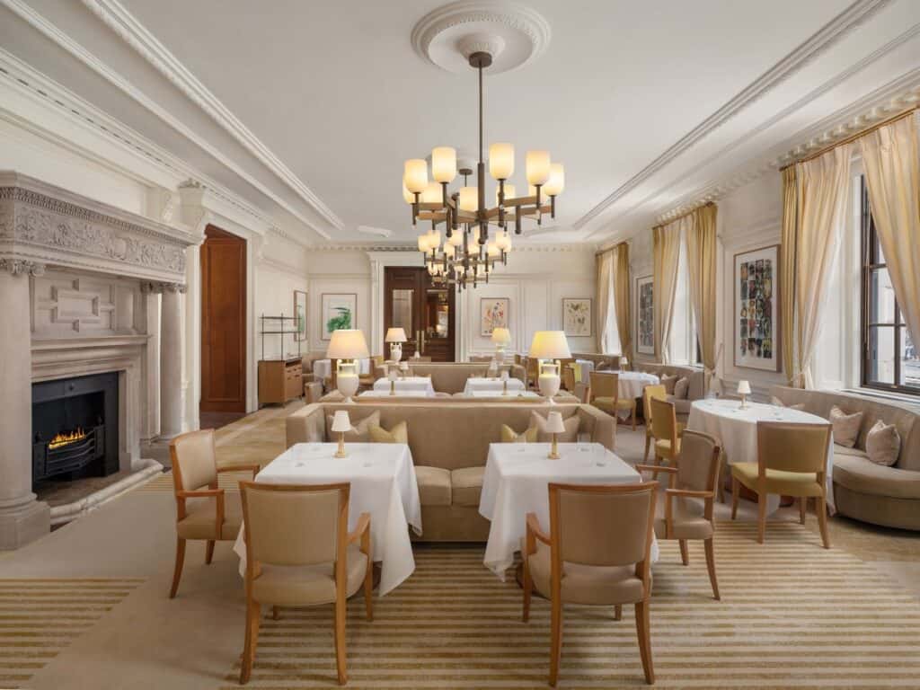 Light-filled dining room at Raffles London with tables in white linen and cream chairs, a carved stone fireplace, tall windows with gold curtains and a tiered chandelier
