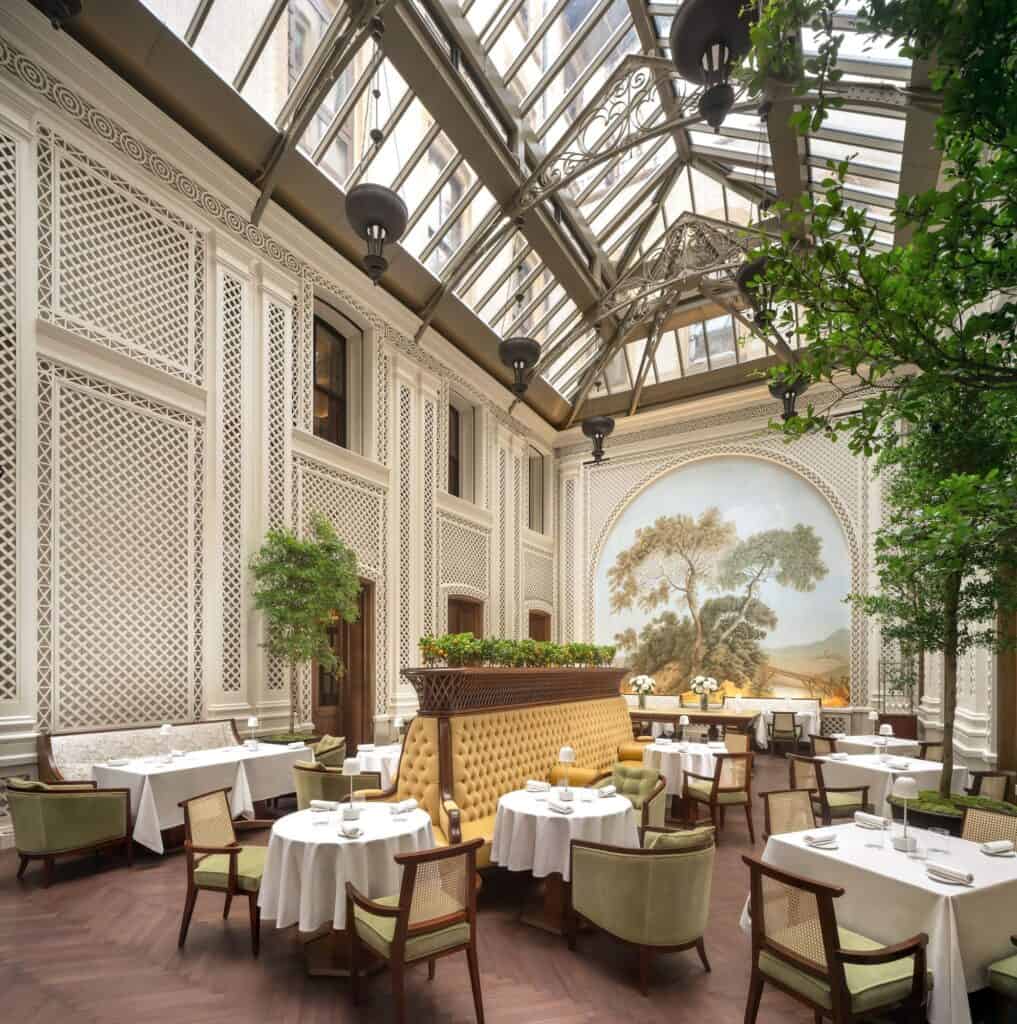 Saison restaurant under a restored glass roof, white lattice-panelled walls, tall indoor trees and green velvet chairs around white tables, facing a painted landscape mural in an arched niche