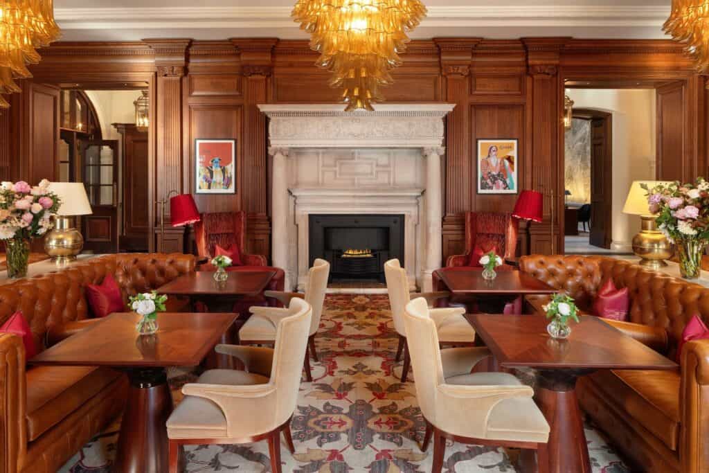 Wood-panelled lounge with tan leather button-back banquettes, cream bouclé chairs and dark timber tables on a patterned carpet, centred on a white marble fireplace beneath amber glass chandeliers