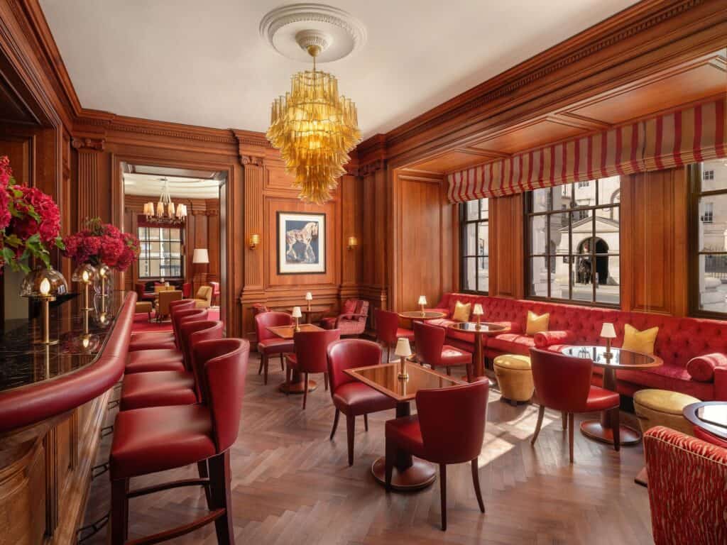 Bar at Raffles London with a curved red leather counter and stools, red banquettes and armchairs, mahogany panelling, a striped red pelmet over the windows and an amber glass chandelier