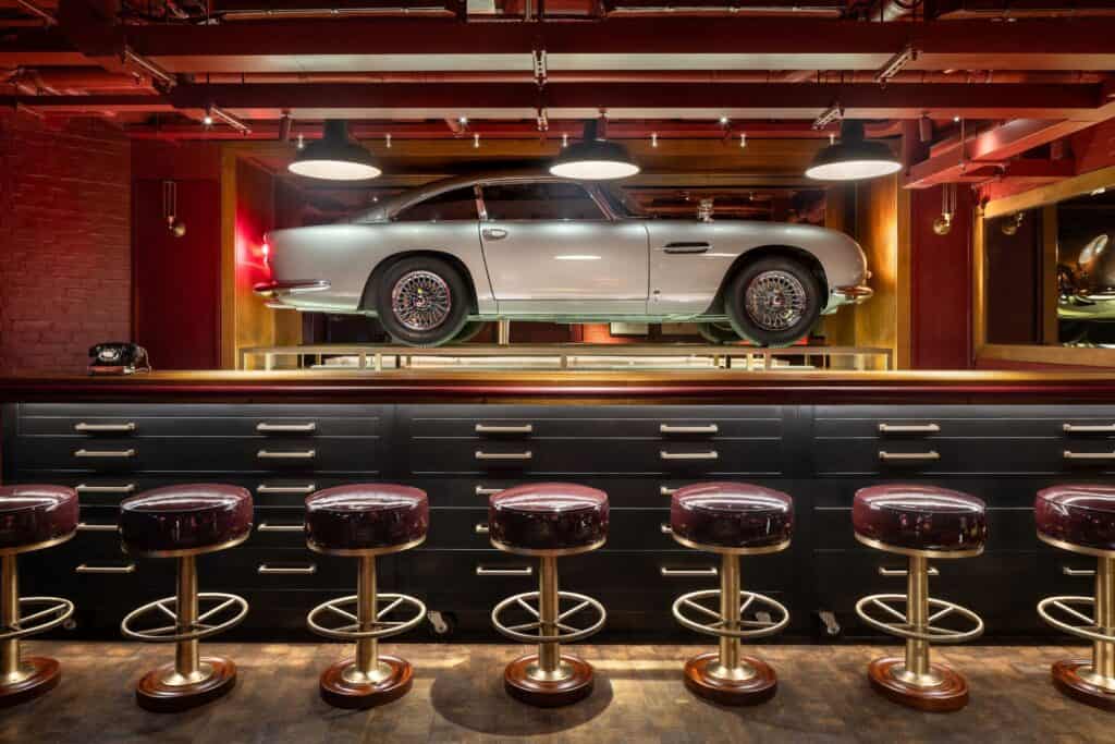 Spy Bar in the basement of Raffles London at The OWO, a silver Aston Martin displayed in a lit recess above a dark bar with a row of burgundy leather stools on brass bases