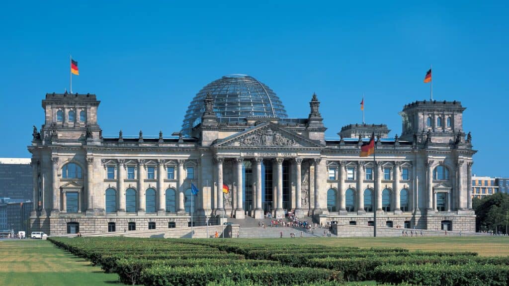 West facade of the Reichstag in Berlin under a clear blue sky, Paul Wallot's colonnaded portico and corner towers crowned by Foster + Partners' glass cupola, German flags flying and a clipped hedge and lawn in the foreground