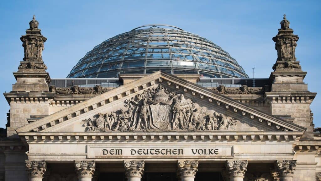 Pediment of the Reichstag carved with the inscription Dem Deutschen Volke and a sculpted coat of arms, flanked by ornate stone towers, with the latticed glass cupola rising behind against a blue sky