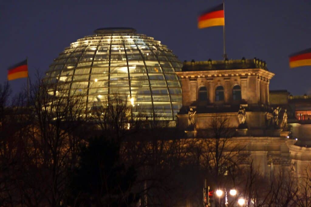 Glass cupola of the Reichstag glowing warmly at dusk behind a sandstone corner tower and bare winter trees, with German flags against a deep blue evening sky