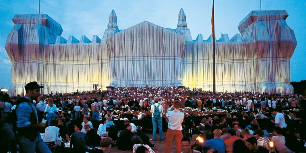 The Reichstag wrapped in silver fabric by Christo and Jeanne-Claude in 1995, its shrouded towers and facade lit at dusk above a dense crowd of visitors on the lawn