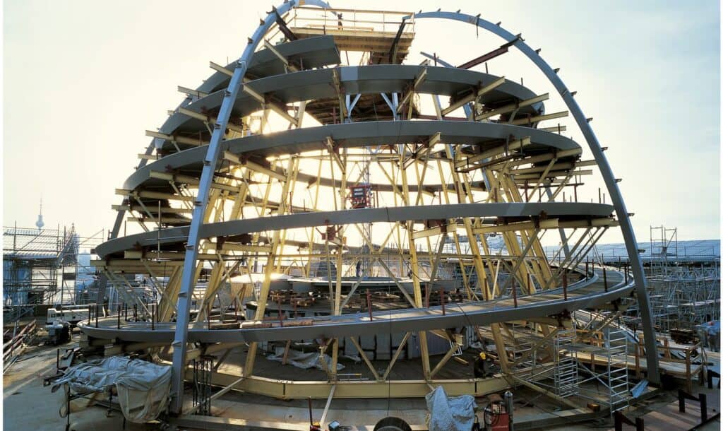 Steel ribs and spiral ramp rings of the Reichstag cupola under construction on the roof, with yellow scaffolding inside and the Berlin TV Tower on the hazy horizon