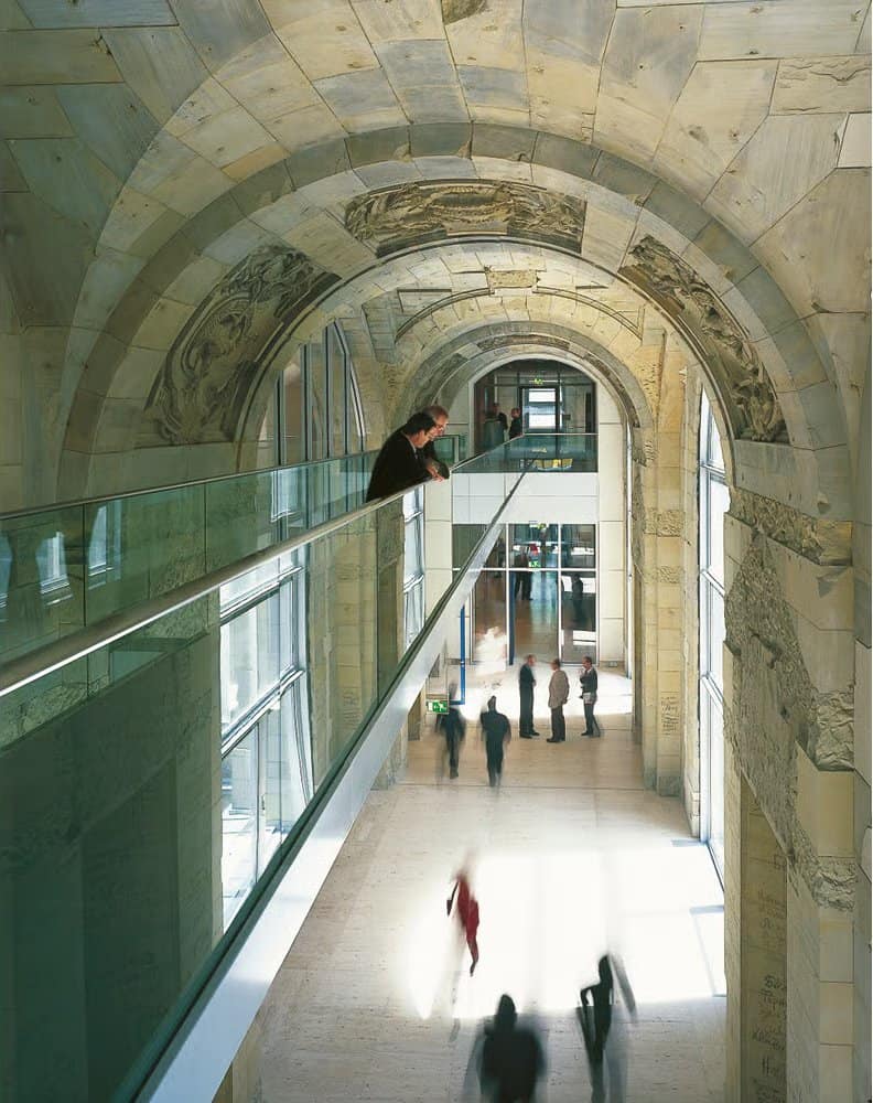 Vaulted sandstone corridor in the Reichstag with carved historic reliefs, a glass balustrade gallery on the left and figures walking along the pale stone floor below