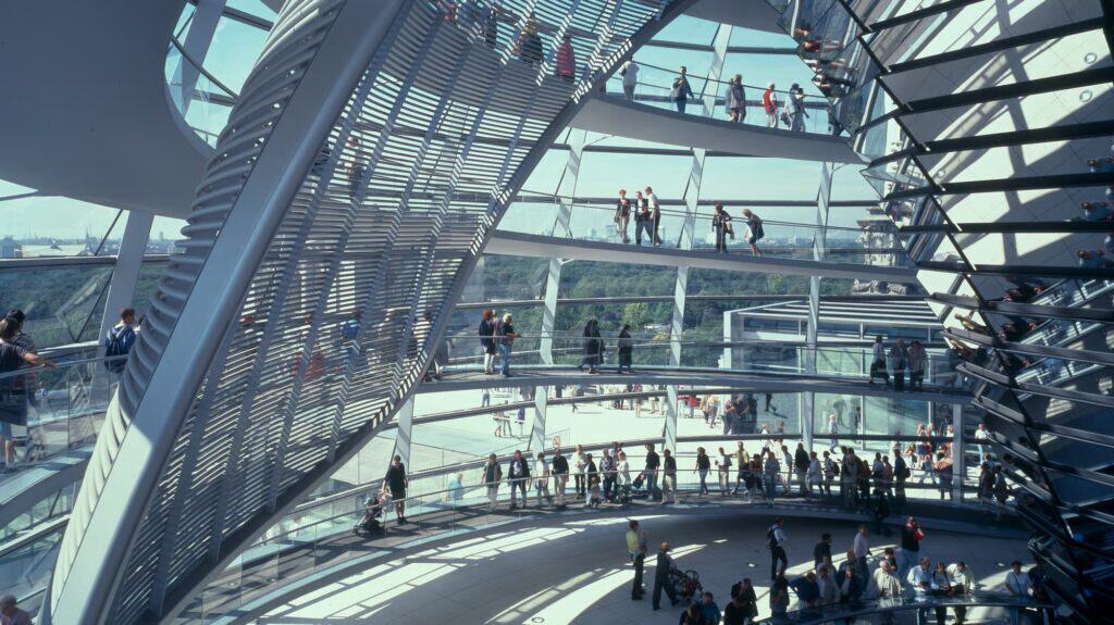 Two spiral ramps sweeping up inside the Reichstag cupola, lined with visitors behind glass balustrades, framed by the slatted sun shield and the steel ribs of the dome